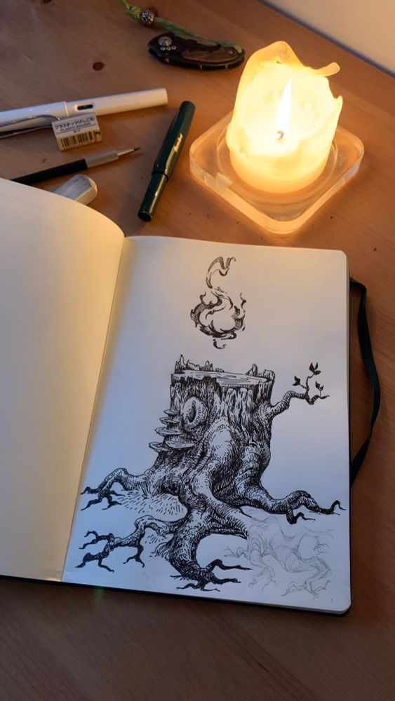 Photo of a sketchbook with a tree with a flame on top. The book lies on a wooden table and a candle lights the scene.