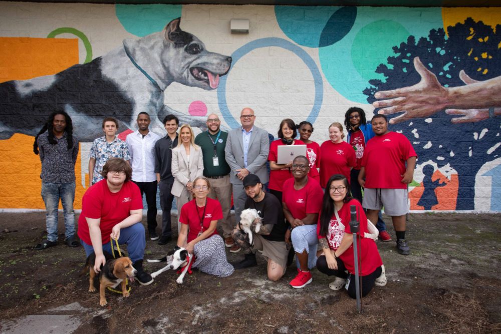 a group of people posing outside of our acps shelter with a mural of a dog behind them