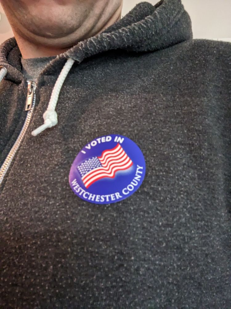 A picture of a sticker that says "I voted" with an American flag on it, stuck to a grey hoodie in a selfie