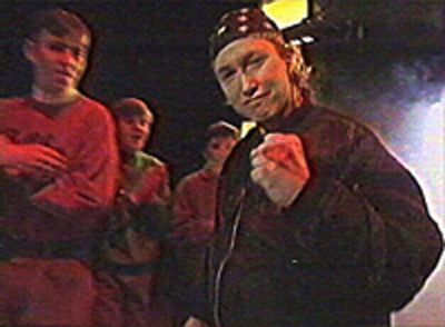 A young gamer called Dave Perry from the 90s. He's wearinga black bomber jacket (as was the style at the time) and a spotted bandana on his head. He's smirking at the camera and clenching his fist triumphantly.