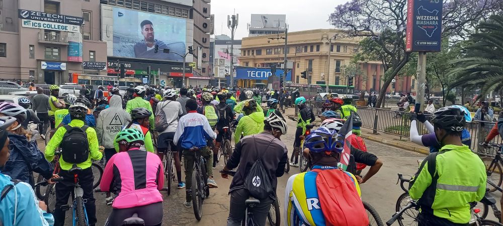 Group of cyclists on a street, mostly with helmets and bike clothes. Grey skies. In the background one older house with colums and another with a large billboard, some trees and buses