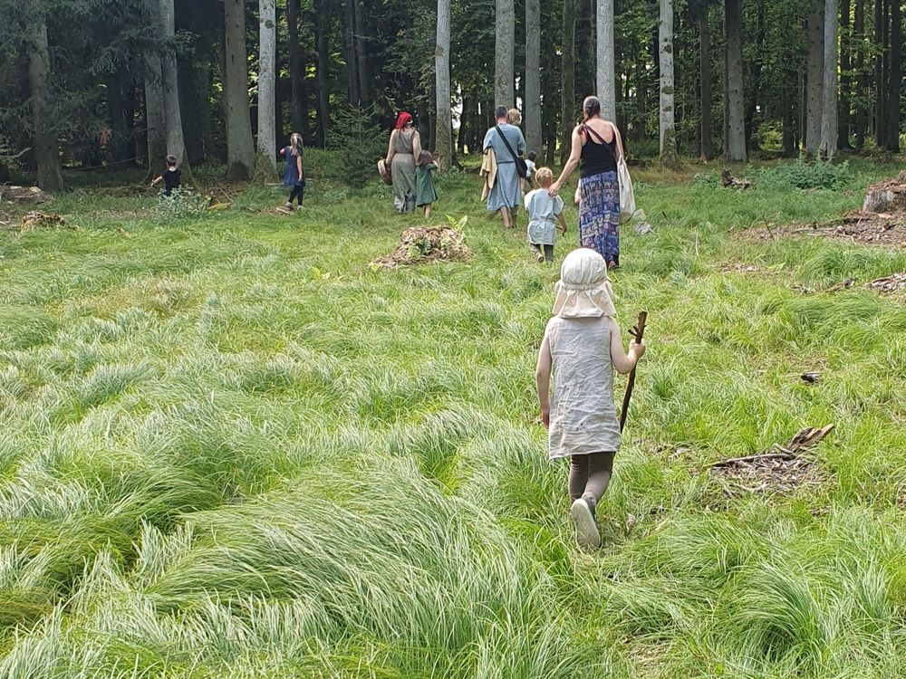 Zezadu focená skupina lidí v generickém fantasy oblečení v lese. V popředí malý chlapec ve dlouhé lněné tunice bez rukávů a s klackem jako holí.