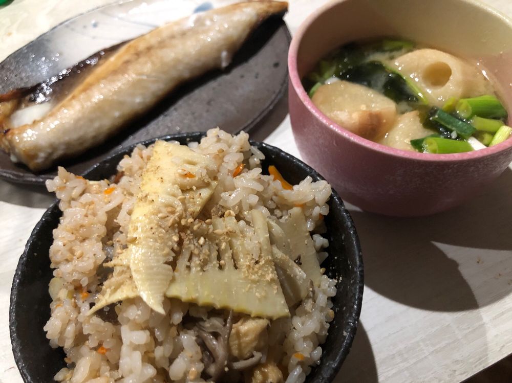 タケノコ2枚が飾られたたけのこご飯、上にはすりゴマがかかっており、黒舞茸、人参、薄揚げと一緒に具だくさん仕様
右上にでっかいお麩とネギとワカメの味噌汁
左上に焼いたサバがお腹を見せてドーン