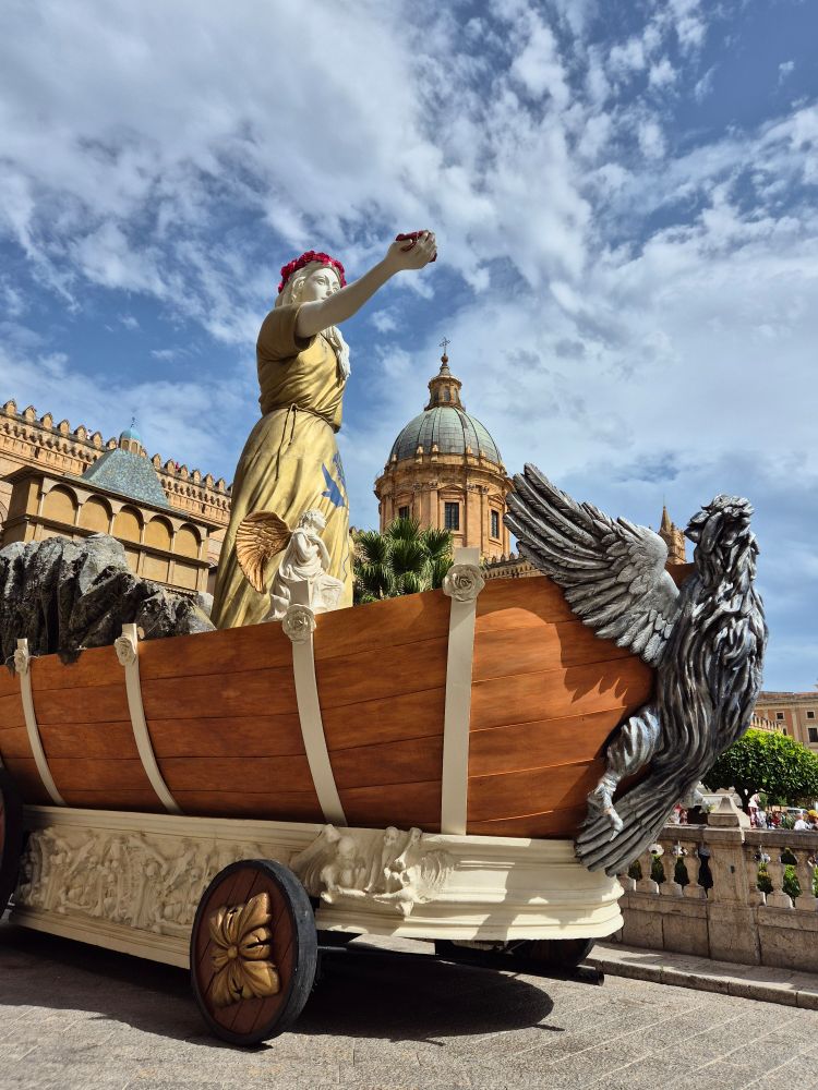 Das Bild zeigt den Festwagen (Carro di Santa Rosalia), der bei den jährlichen Feierlichkeiten zu Ehren der Heiligen Rosalia in Palermo, Sizilien, verwendet wird. Eine kurze Bildbeschreibung:
Ein prunkvoller Festwagen mit einer goldfarbenen Statue einer Frau und großen silbernen Flügeln steht vor einer historischen Kuppelkirche bei blauem Himmel mit Wolken.

