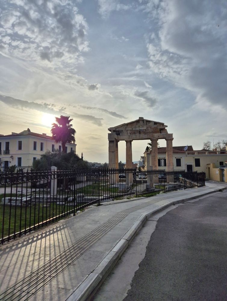 Das Bild zeigt eine antike Tempelruine mit Säulen in Athen, Griechenland. Im Hintergrund ist ein Sonnenuntergang zu sehen, der die Szene in warmes Licht taucht. Umgeben ist die Ruine von einem schwarzen Metallzaun, und rechts führt eine gepflasterte Straße vorbei. 