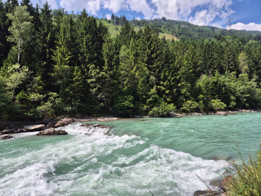 Ein türkisfarbener Fluss fließt mit leichtem Wildwasser durch eine bewaldete Berglandschaft unter blauem Himmel mit einigen Wolken. 