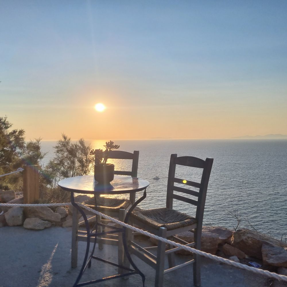 Ein kleiner Tisch mit zwei Stühlen steht abgesperrt hinter einem Seil auf einer Terrasse mit Blick aufs Meer, während die Sonne tief über dem Horizont untergeht. 