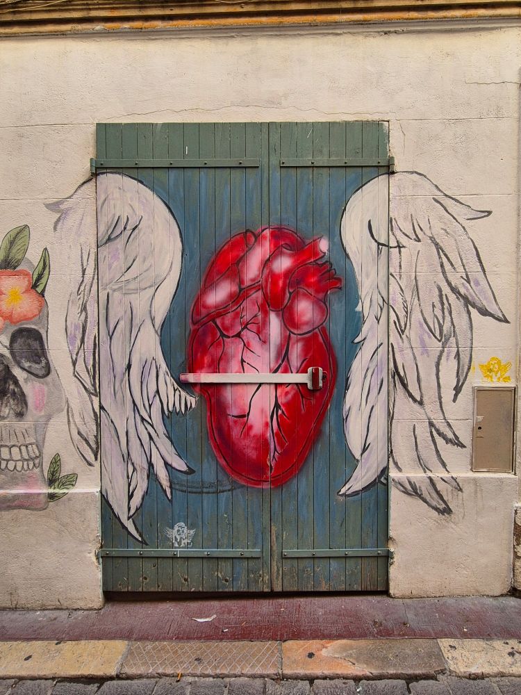 Ein großes rotes Herz mit weißen Engelsflügeln, gemalt auf grünen Holzläden einer Tür. Links erkennt man einen Totenkopf mit Blumen.