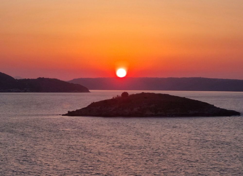 Ein kleiner, bewaldeter Hügel auf einer Insel im Meer wird von einem leuchtend orange-roten Sonnenuntergang hinterleuchtet. Die Sonne steht tief über dem Horizont und taucht Himmel und Wasser in warme Farben. 