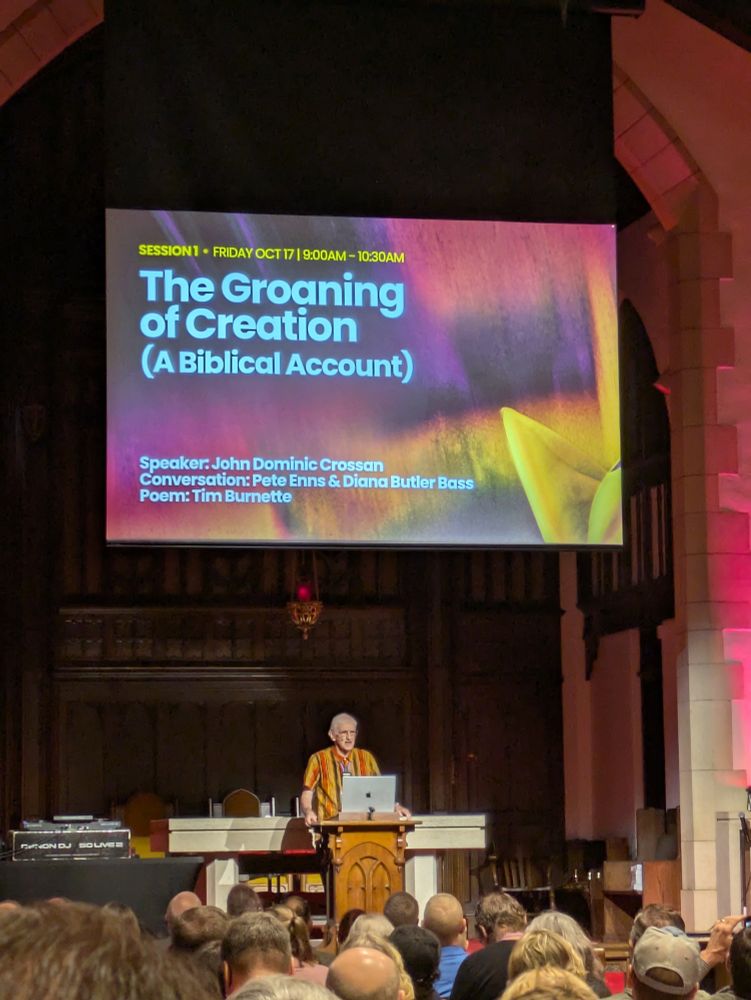 John Dominic Crossan stands at a podium in a cathedral-like church, delivering a presentation titled “The Groaning of Creation (A Biblical Account)”. He wears a vibrant, multicolored robe and addresses an attentive audience seated in pews. Behind him, a large screen displays session details: Session 1 – Friday Oct 17 | 9:00AM–10:30AM, with featured contributors Pete Enns, Diana Butler Bass, and poet Tim Burnette.