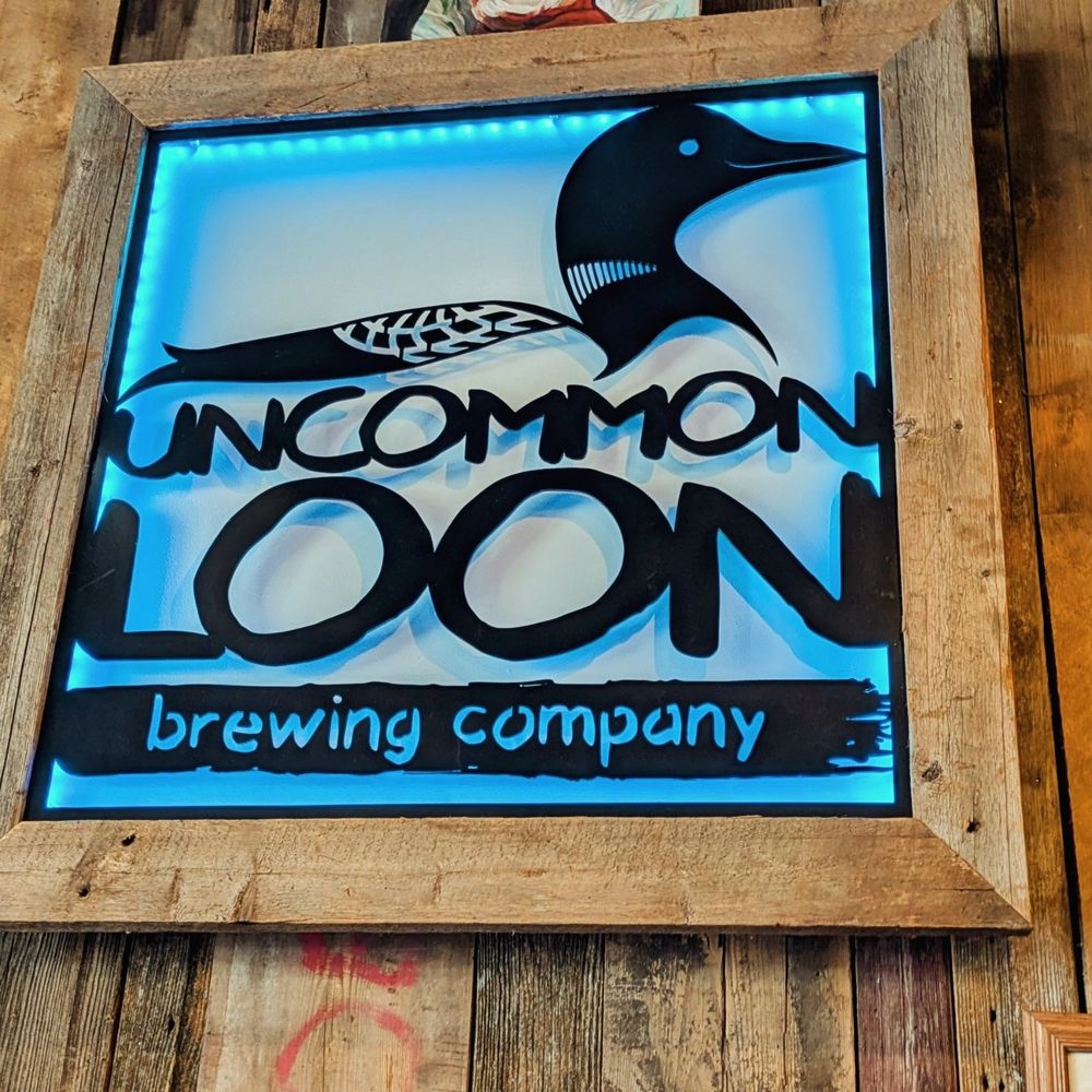 Wooden sign for Uncommon Loon Brewing Company, featuring a stylized loon bird and blue backlighting. The sign is framed in rustic wood with a textured plank background, evoking a cozy, Northwoods aesthetic.