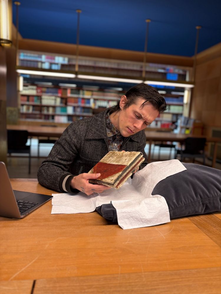 Mitchel studying the manuscript in the reading room. In front of him is a book pillow and Tyvek sheets. On the right is his laptop. In the background you see the modern reference library.