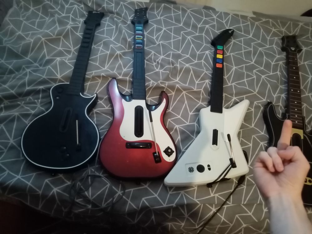 a display of my guitar hero guitar collection. from left to right: a modded 360 les paul, a guitar hero 5 360 guitar, a guitar hero 2 xplorer and, slightly off screen with me visibly flipping it off, a guitar hero live guitar 