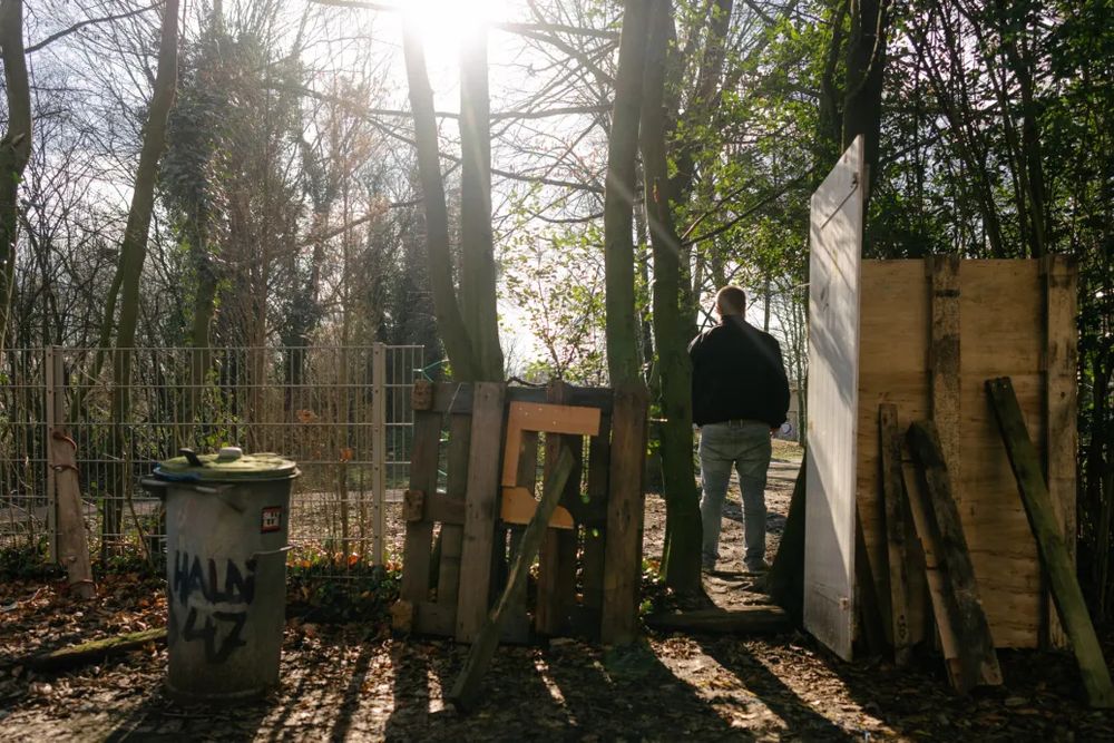 Mann, mit Rücken zur Kamera steht in einem zurechtgebastelten Gartentor. Links steht ein metallener Mülleimer mit dem Tag "haldi 47"