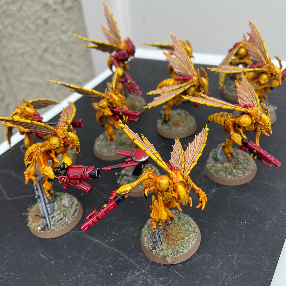 Ten painted T’au Vespid Stingwing miniatures with orange-yellow bodies, bright red guns, and pale cream wings with brown shading. An additional Oversight Drone is present, painted in matching colours. All models are on round bases with sand, rocks, and tufts of green and brown grass.