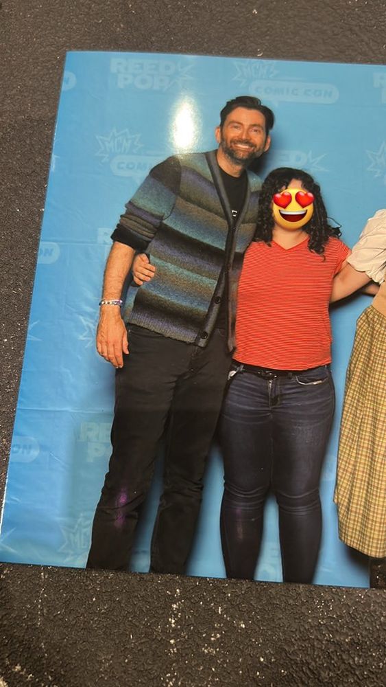 Girl in red shirt and jeans is with David Tennant’s who is in black pants and blue sweater and she has her arm wrapped around his waist.