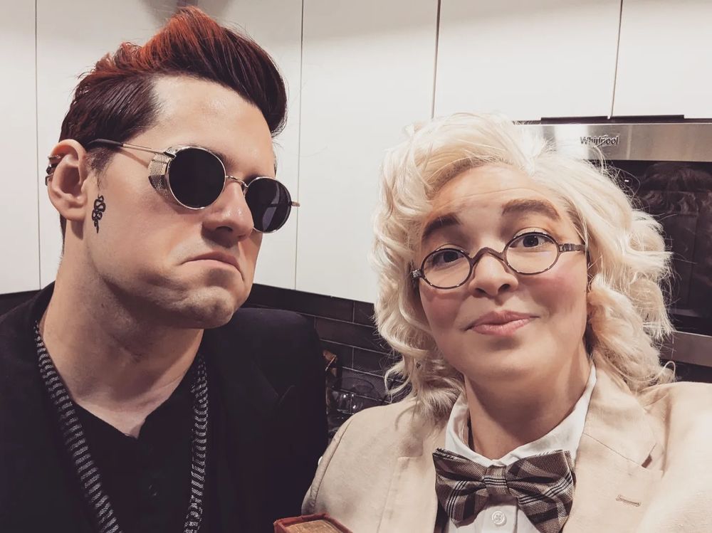 Close up of Jeff and Lana as Crowley and Aziraphale, respectively. Crowley is still drowning and there is a small snake tattooed in a cascade beneath his right sideburn. Aziraphale looks pleased at the simple human pleasure of a photo with her best friend.