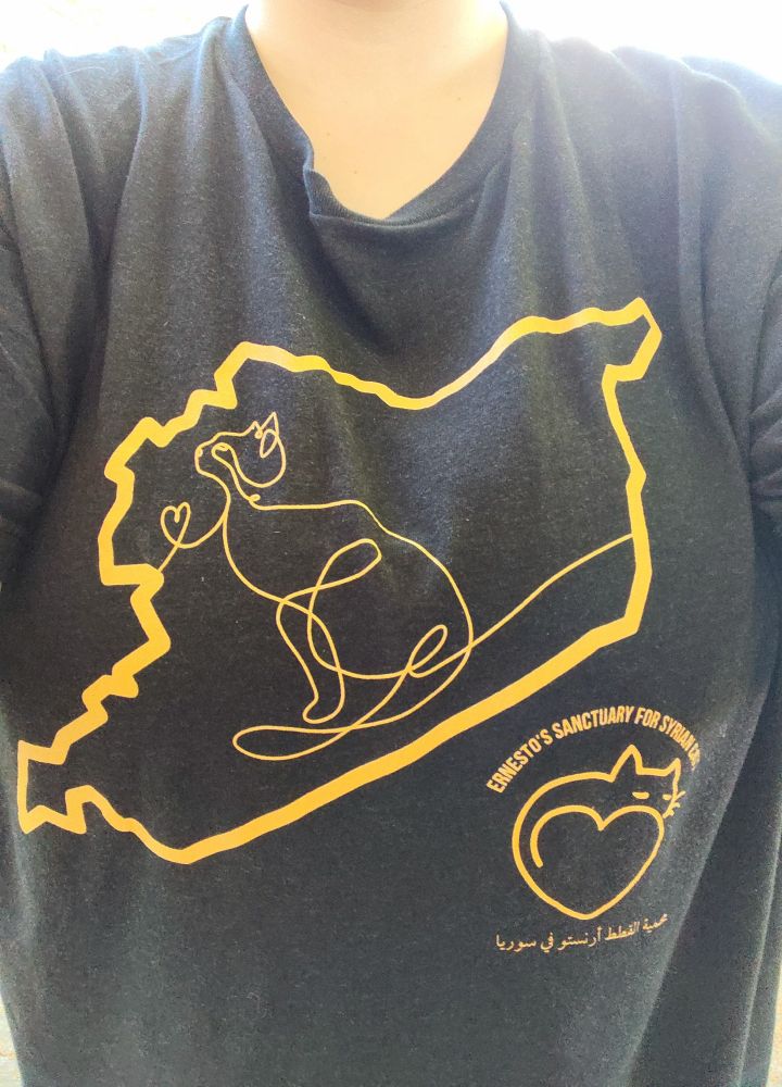 Black tee shirt with golden design of an outline of Syria, featuring line art of a curious cat and in the middle. On the right side under Syria is the Ernesto's logo with the text "Ernesto's Sanctuary for Syrian Cats" in English and Arabic.