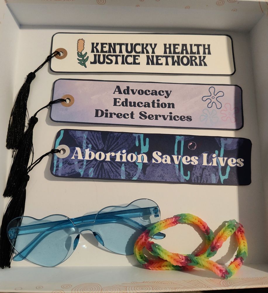 White box displaying three bookmarks made out of cardstock, with black tassels on the top side. The bookmarks are: White background with The Kentucky Health Justice Logo; a purple coral/floral background with black text saying "Advocacy, Education, Direct Services"; a blue/purple floral wave background with white text saying "Abortion Saves Lives"