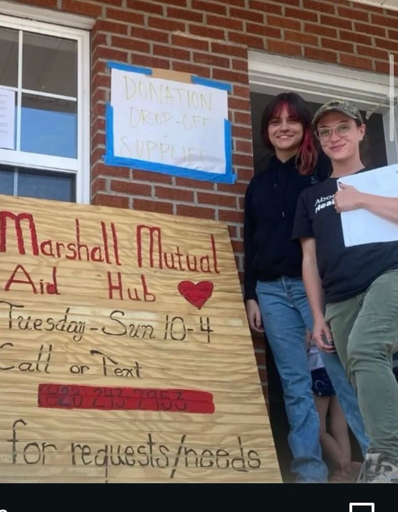 Two people stand beside a sign that reads “Marshall Mutual Aid Hub” after Hurricane Helene in WNC.