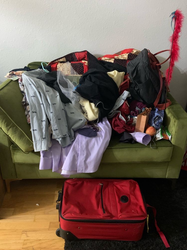 A pile of clothes on a green couch. It is a big pile of clothes that will probably not fit in the small carry on suitcase.