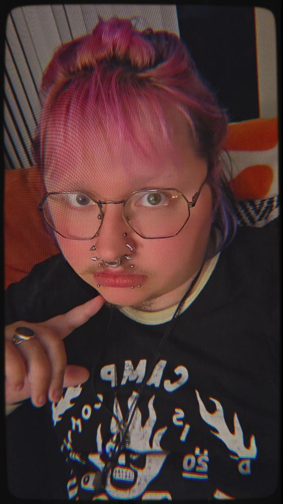 Kit (he/they/it/xe) with pink, purple, and black hair; multiple facial piercings; and a Camp Unus Annus shirt on. 