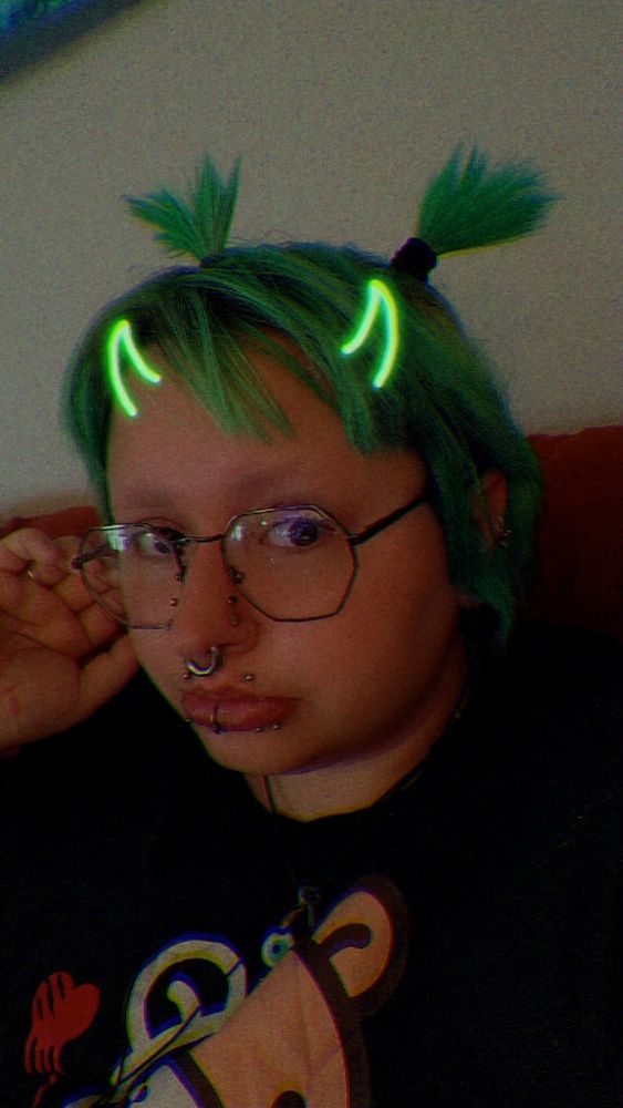 Kit (he/they/xe/it) with green hair in pigtails, green horns (filter), and multiple facial piercings. 
