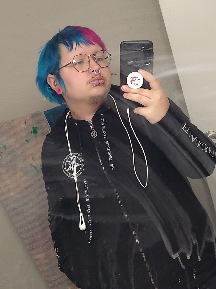 Kit (he/they/it/xe) with blue and pink split dyed hair; pink, blue, and silver piercing jewelry, and a Black Craft hoodie.