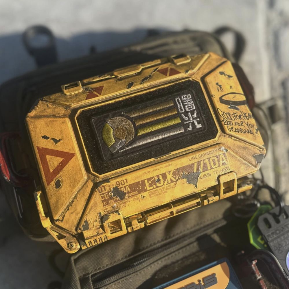 An angular yellow industrial-looking hardcase has been attached to a drab green chest pouch. The case has red arrow markings on two sides and a supply kit label on the bottom. It has been defaced with graffiti which reads 'GOOD FOR HEALTH BAD FOR YO GRANDMA' and it battered and grimy. There is a patch affixed to the case depicting an astronaut's helmet in profile.