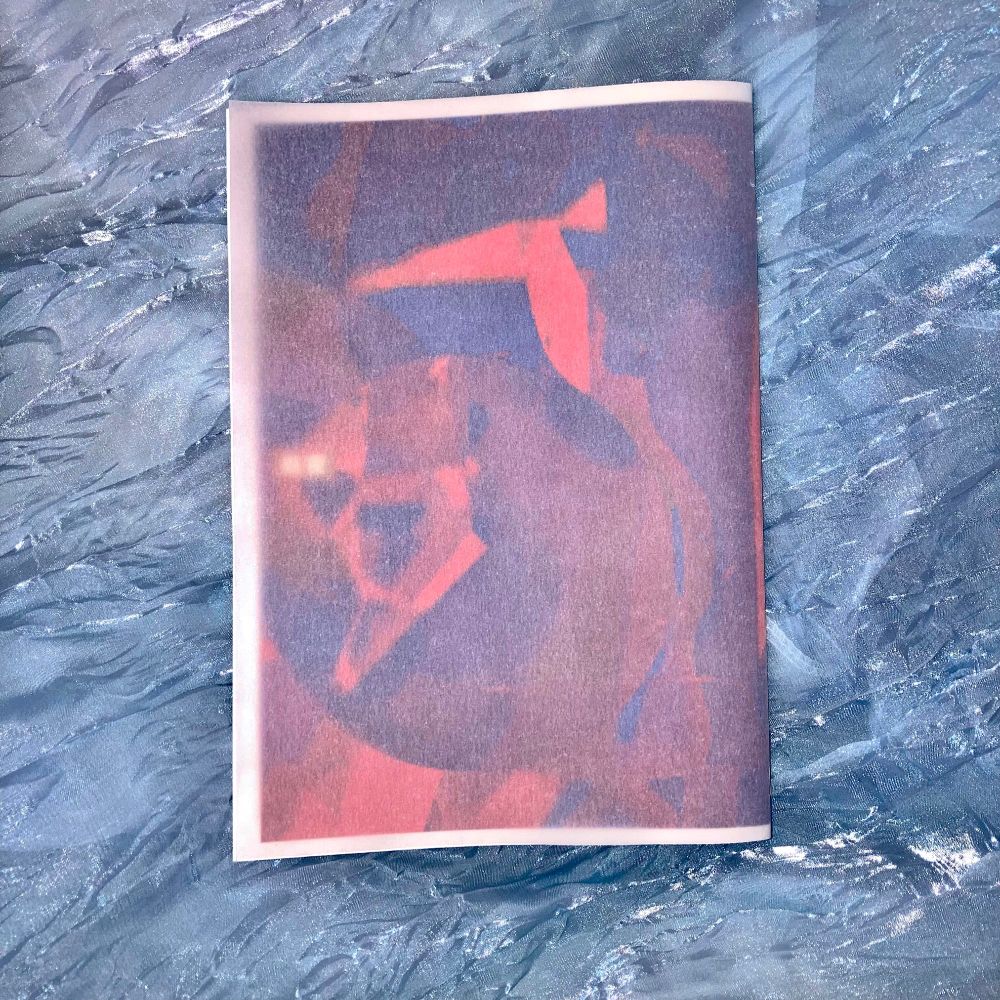 ネットプリントで作った冊子「前夜」の写真。薄い青色の、波打っているような素材の生地の上に冊子を乗せている。2枚目の写真に写っていた鮮やかな色合いの表紙の上から白色のトレーシングペーパーをつけたバージョン。色味が穏やかになっている。