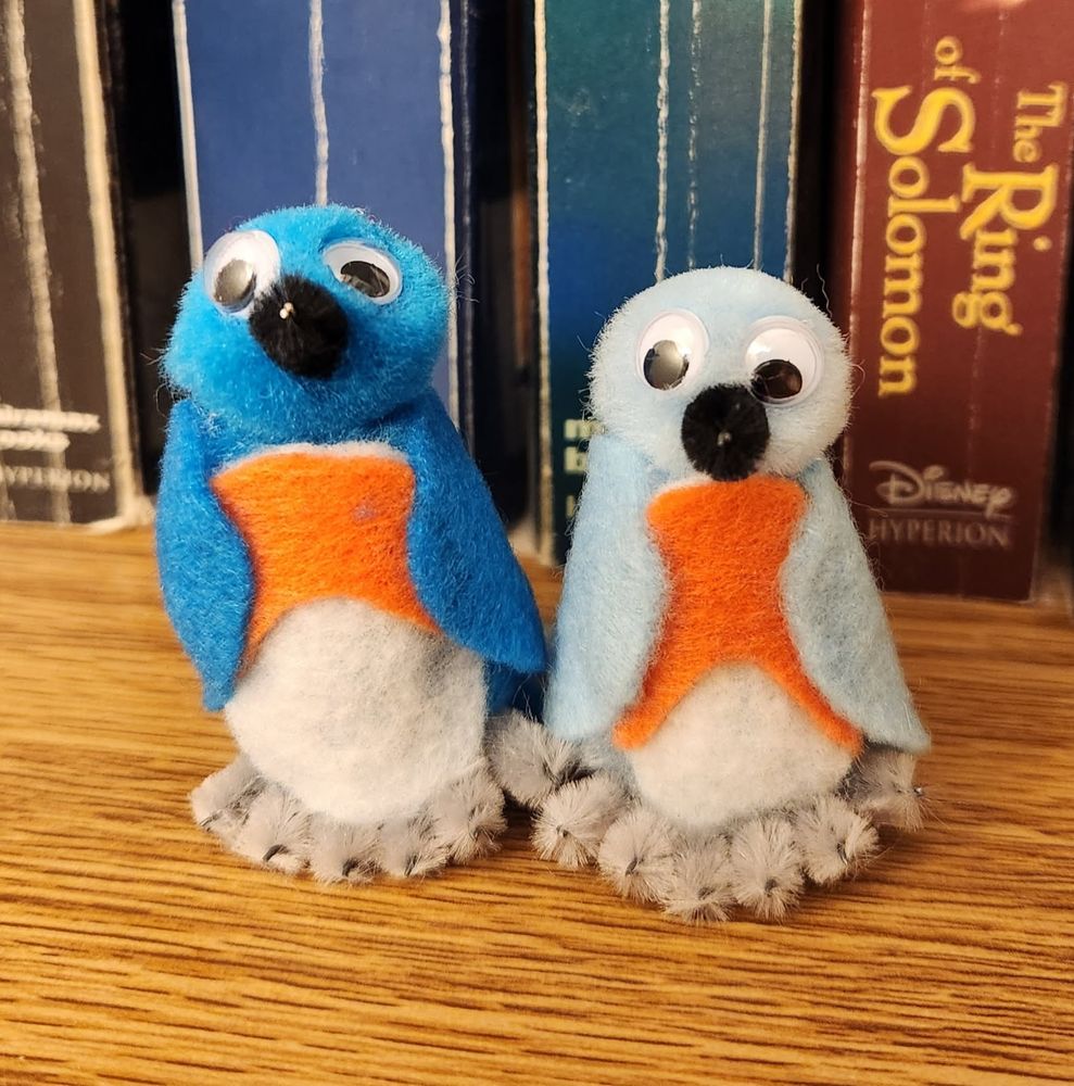 A handcrafted pair of Eastern Bluebirds sitting side by side on a wooden bookshelf, facing directly toward the camera. The birds' bodies and heads are made of blue pompoms: bright blue for the male on the left and paler blue for the female on the right. Their wings, tucked against their sides, are made of blue felt. They have a white belly and orange chest, both felt. Their feet are made of light gray pipe cleaners. They each have a black pipe cleaner beak and a pair of googly eyes. The bluebirds are each about 2 inches tall.