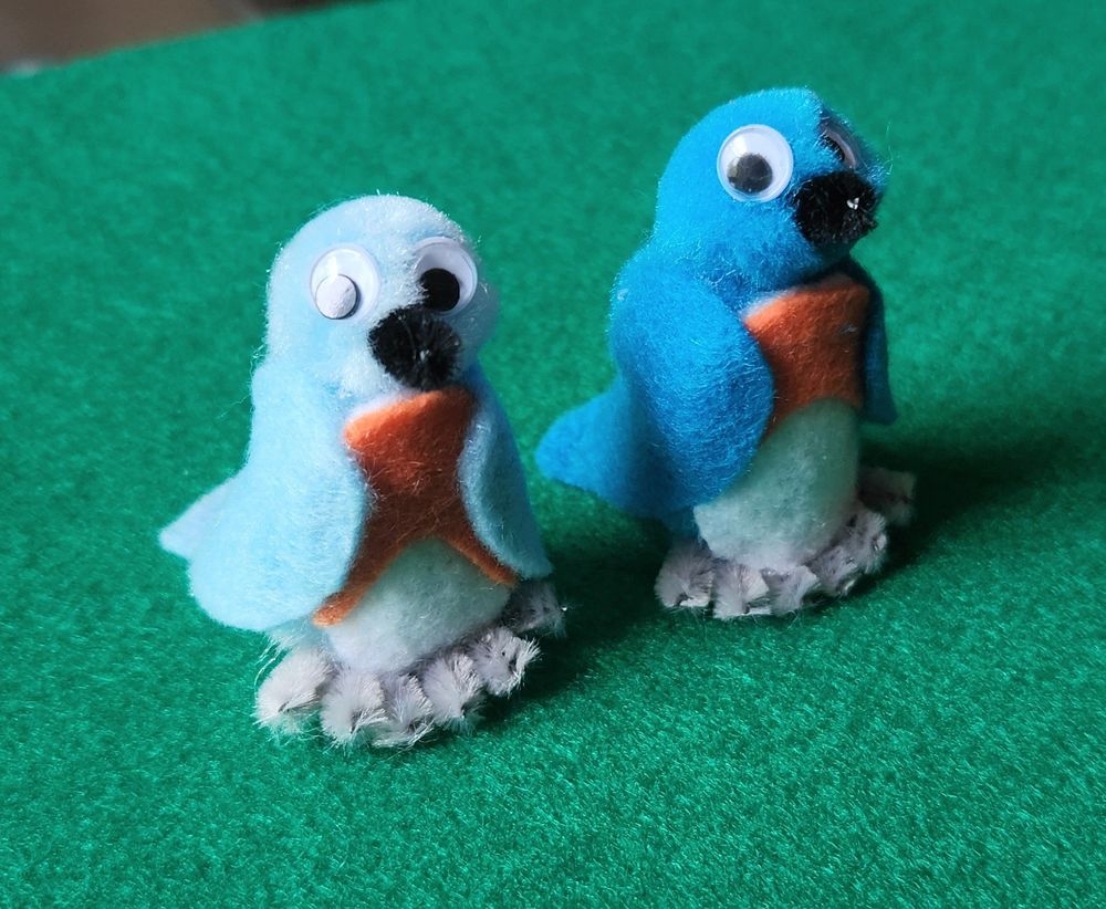 A handcrafted pair of Eastern Bluebirds sitting side by side on a green felt background, facing partly to the right and partly toward the camera. The birds' bodies and heads are made of blue pompoms: bright blue for the male on the right and paler blue for the female on the left. Their wings, tucked against their sides, are made of blue felt. They have a white belly and orange chest, both felt. Their feet are made of light gray pipe cleaners. Their tails, only partially visible behind the birds, are made of blue felt and rest against the ground, providing balance and support. They each have a black pipe cleaner beak and a pair of googly eyes. The bluebirds are each about 2 inches tall.