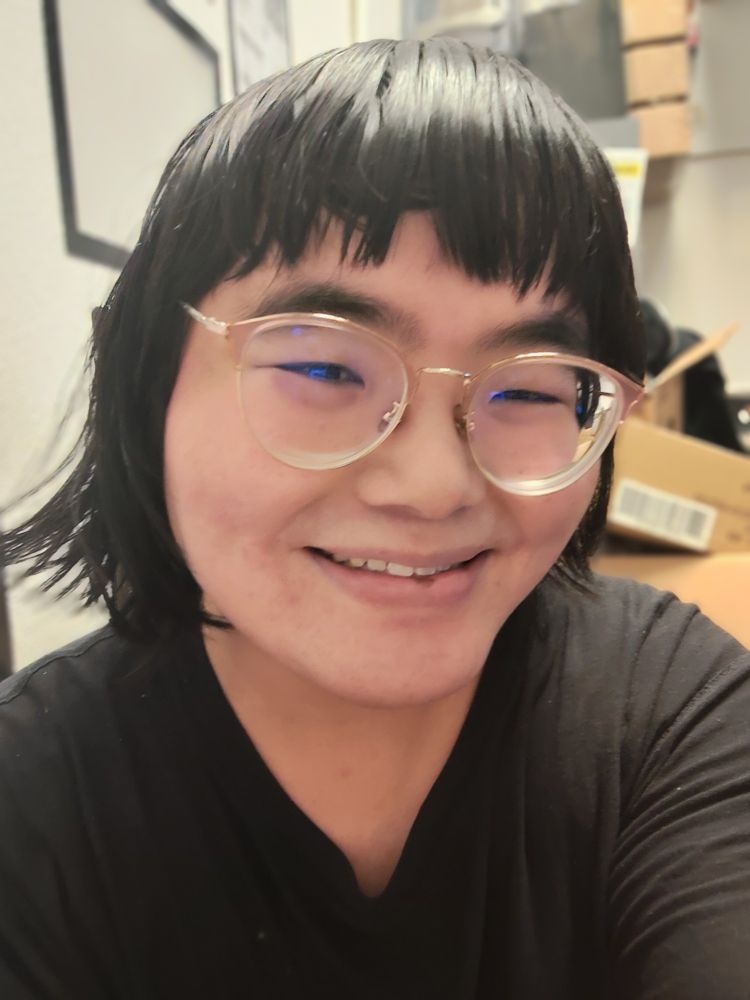 Selfie of an east Asian trans woman with short hair and glasses smiling 