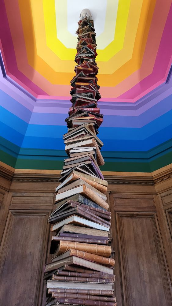 An art installation of books stacked leads to a colorful ceiling and a statue of a child is perched on top.