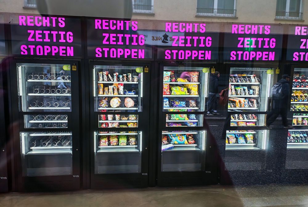 Blick in einen 24h-Automatenkiosk. Auf der LED-Anzeige über den Automaten steht "Rechts zeitig stoppen".