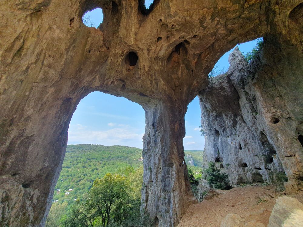 Blick durch mehrere Felsbögen hinunter auf bewaldete Hügel. In der Decke der Felsformation ist der Himmel durch mehrere Löcher zu sehen.