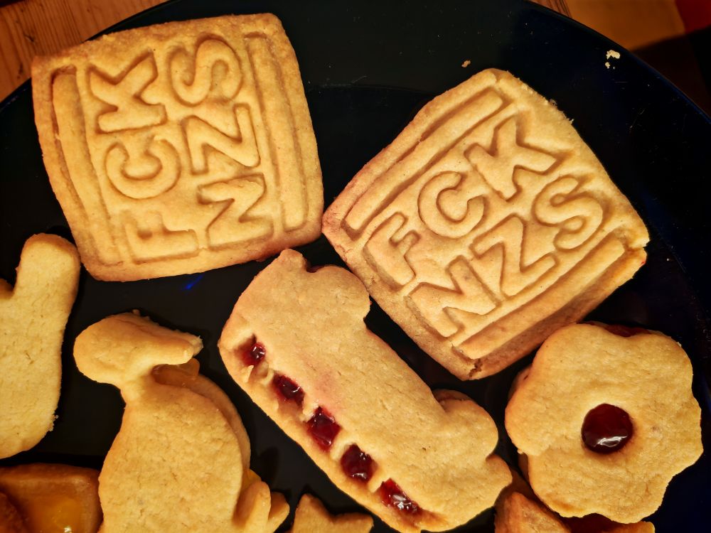 Plätzchen von oben fotografiert. Zwei quadratische Plätzchen, in die die Aufschrift "FCK NZS" eingeprägt ist, ein Van (sieht aus wie ein alter VW-Bus), bei dem rote Marmelade aus den Fenstern guckt, ein Blume mit roter Marmelade in der Mitte, eine Ente.