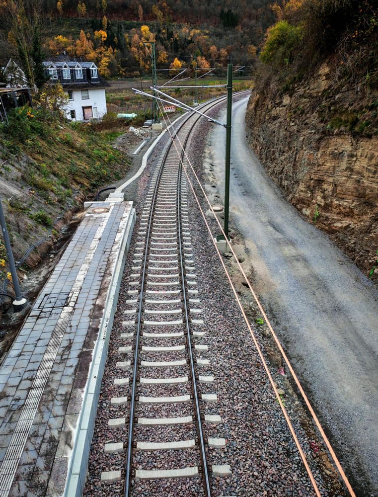 Blick von der gleichen Stelle über die Oberleitungen und die Bahnstrecke, diesmal der Blick die Gleise entlang. Rechts und links der Bahnstrecke sind steile, in Herbsttönen bewachsene Hänge. Links steht im Hintergrund ein weißes Gebäude.