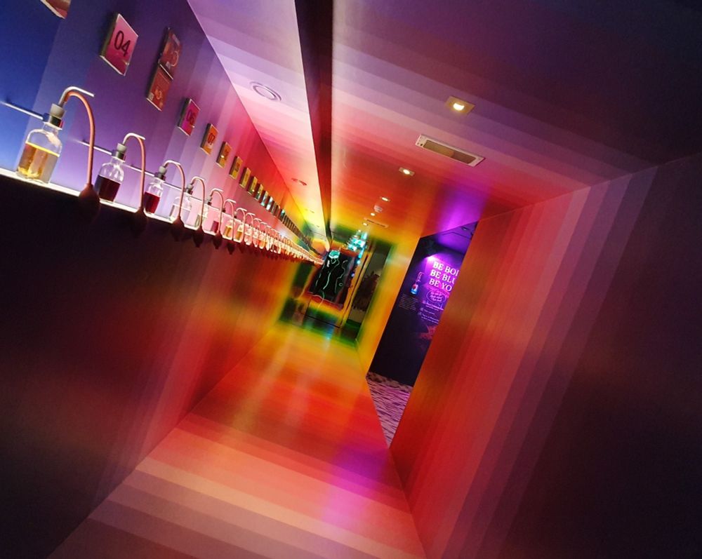 Blick durch einen Flur, dessen Boden, Wände und Decke in farbigen Streifen von lila über rot zu orange, gelb und grün gehalten sind. Am Ende des Flurs sorgt ein Spiegel für eine optische Verlängerung.
Auf der linken Seite stehen Fläschchen mit verschiedenfarbigen Flüssigkeiten mit Schläuchen darin auf einem Absatz den Flur entlang. Hinten rechts ist ein Durchgang, in dem man lila Licht sieht. Das Foto ist in einem Winkel von etwa 30 Grad geneigt. 
