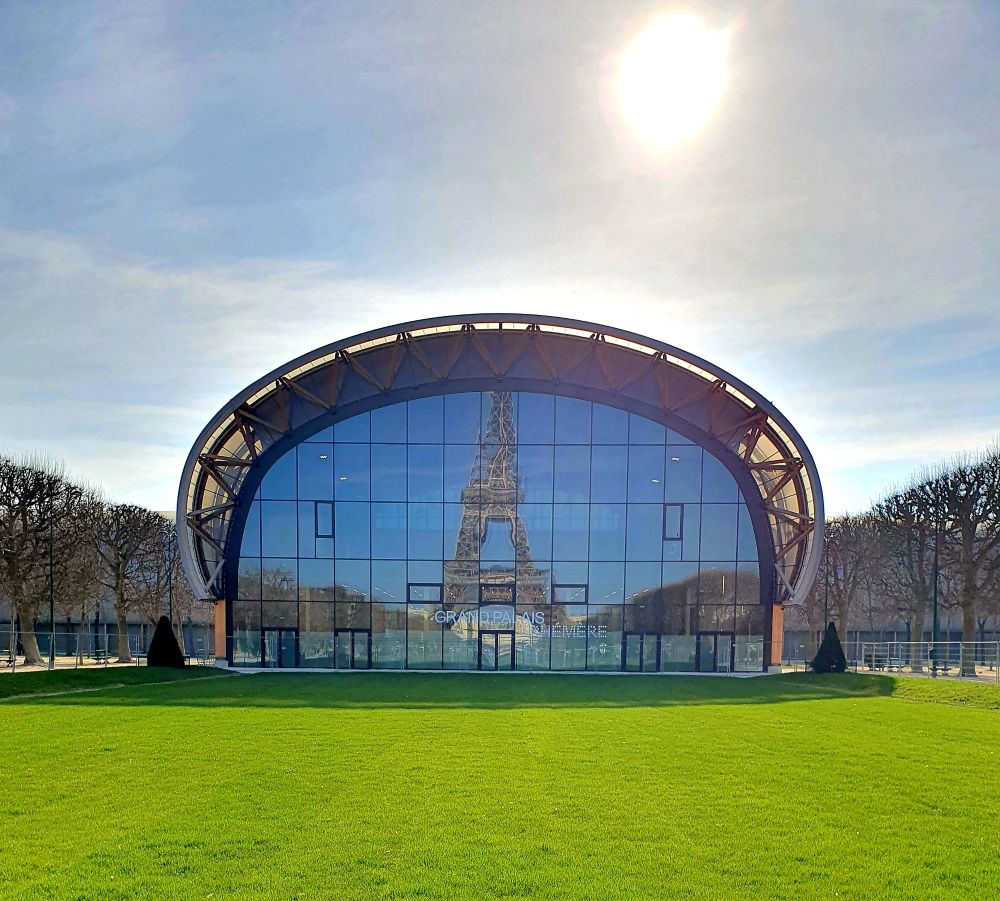 Auf einem leuchtend grünen Rasen steht ein halbrundes großes Gebäude, was nach vorne eine gerade spiegelnde Fensterfront hat. In der Fensterfront spiegelt sich der Eiffelturm vor blauem Himmel. Über dem Gebäude steht rechts eine gleißende Sonne.