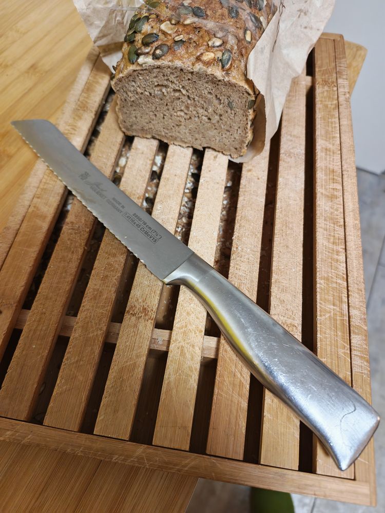 Hölzerner Rost auf einer Art hölzernem Tablett, dessen Umrandung rund um den Rost reicht. Darauf liegt ein angeschnittenes Brot und ein Brotmesser.