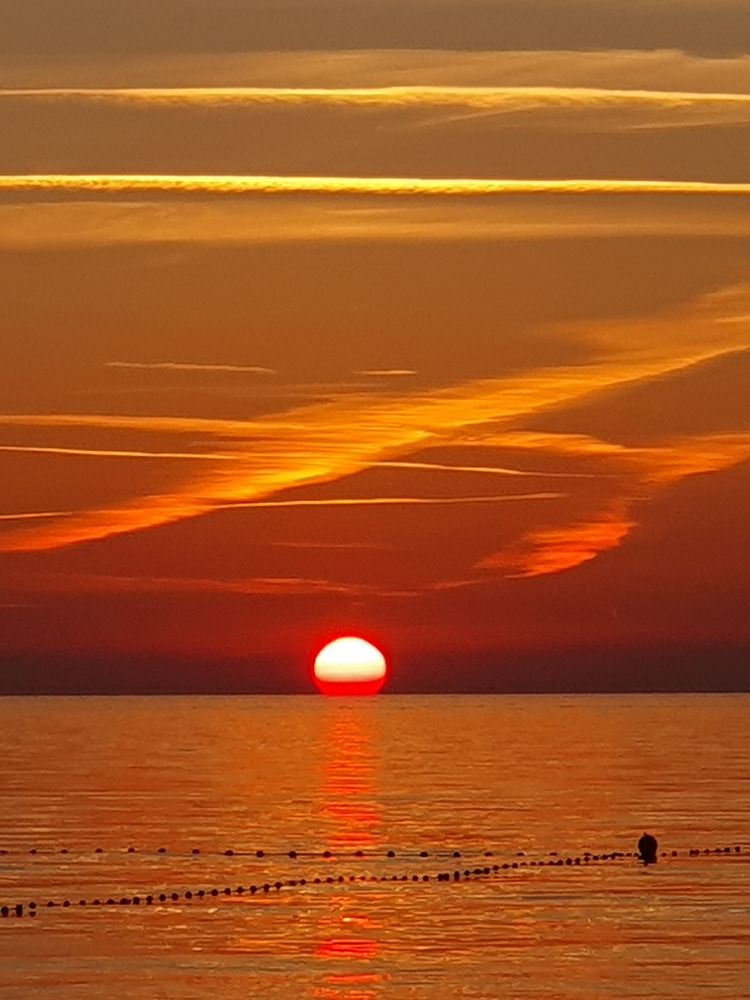 Sonnenuntergang. Die Sonne ist schon ein kleines Stück hinterm Meer verschwunden und leuchtet orange. Meer und Himmel sind sehr orange gefärbt. 
