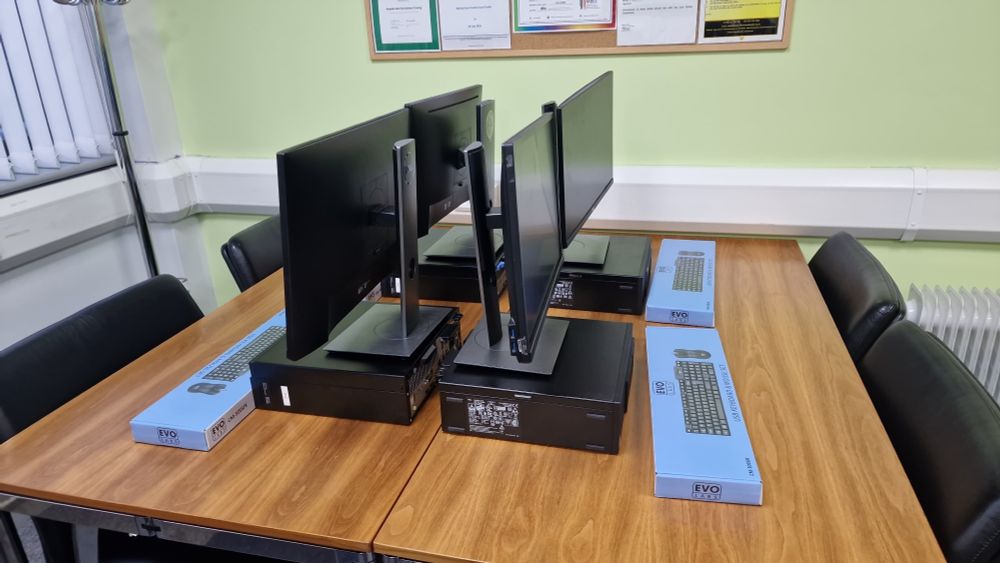 Four new table top computers with screens and keyboards on a table with a chair facing each computer and screen them ready for use.