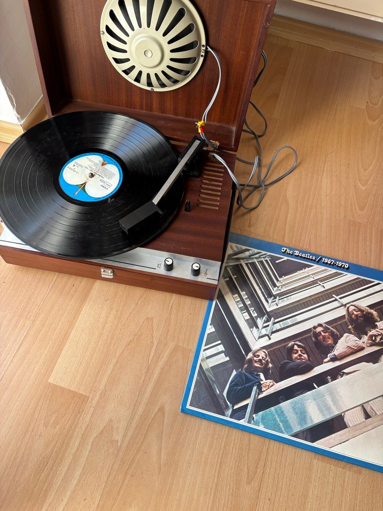Ein Vintage-Plattenspieler mit Holzgehäuse spielt eine Schallplatte von The Beatles. Das Album ‘The Beatles 1967–1970’ mit blauem Cover liegt daneben auf dem Holzboden. Der Plattenspieler hat einen eingebauten Lautsprecher im geöffneten Deckel, und der Tonarm ist auf der Schallplatte positioniert. Ein paar Kabel sind sichtbar, und das Gerät zeigt leichte Gebrauchsspuren.
