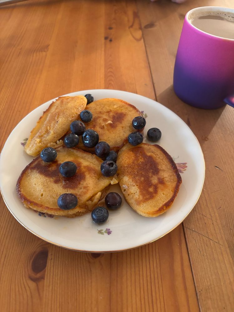 Ein Teller mit kleinen, goldbraun gebratenen Pfannküchlein, die mit frischen Blaubeeren garniert sind, steht auf einem alten Holztisch. Die Pfannküchlein haben eine knusprige Kruste und eine weiche, fluffige Mitte. Der Teller ist weiß mit zartem Blumenmuster und hat einen goldenen Rand. Daneben steht eine Tasse mit Kaffee, deren Farbverlauf von Lila zu Pink übergeht. Eine gemütliche Frühstücksatmosphäre.