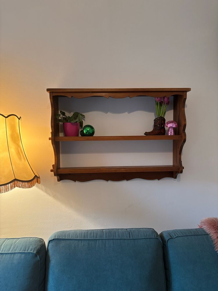 Ein gemütlicher Wohnbereich mit einer Vintage-Atmosphäre. An der Wand hängt ein hölzernes Regal mit geschwungenen Kanten, das teilweise dekoriert ist. Auf der linken Seite steht eine grüne Pflanze in einem pinken Topf, daneben liegt eine grüne, schimmernde Discokugel. Rechts im Regal befindet sich ein kleiner Cowboystiefel, aus dem frische, pinkfarbeneTulpen herausragen. Daneben steht eine rosafarbene Discokugel in Pilzform.
Unter dem Regal steht ein blaugrünes Sofa aus weichem Cordstoff. An der rechten Seite des Sofas lugt ein rosafarbenes Schafsfell hervor. Links im Bild verbreitet eine klassische Stehlampe mit gelblichem Licht eine warme, wohnliche Stimmung.