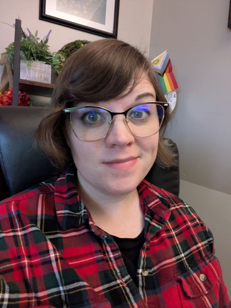 Picture of me sitting in an office chair with office decor behind me. I have short brown hair and black and gold rimmed glasses. I'm wearing a red and green plaid shirt with white and yellow stripes.