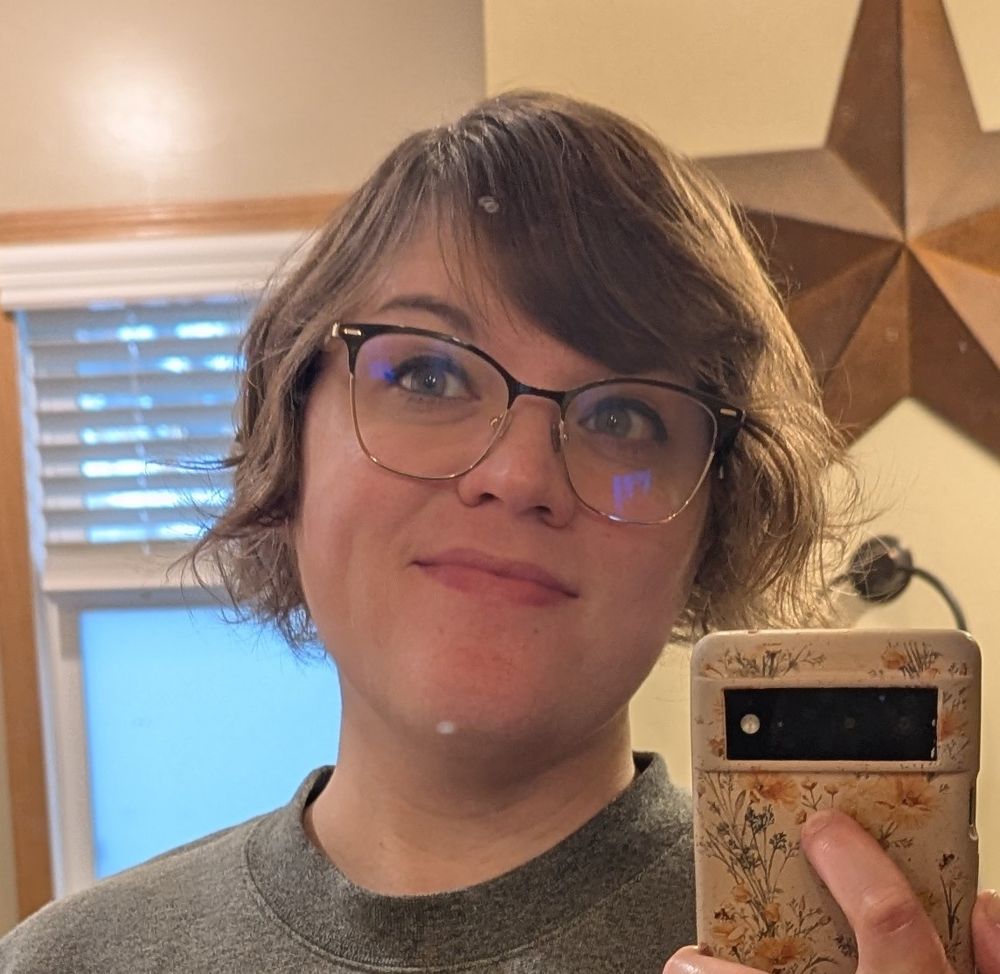 Picture of me taking a selfie in the bathroom mirror. I have short brown hair and black and gold rimmed glasses.