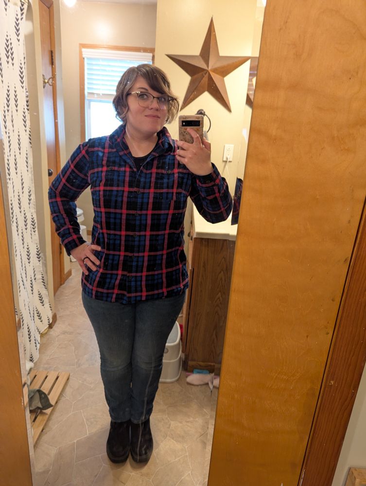 Picture of me taking a photo of my reflection in the bathroom mirror. I'm wearing a black, blue, and pink plaid shirt with blue jeans and black boots. I have short brown hair and black and gold rimmed glasses.