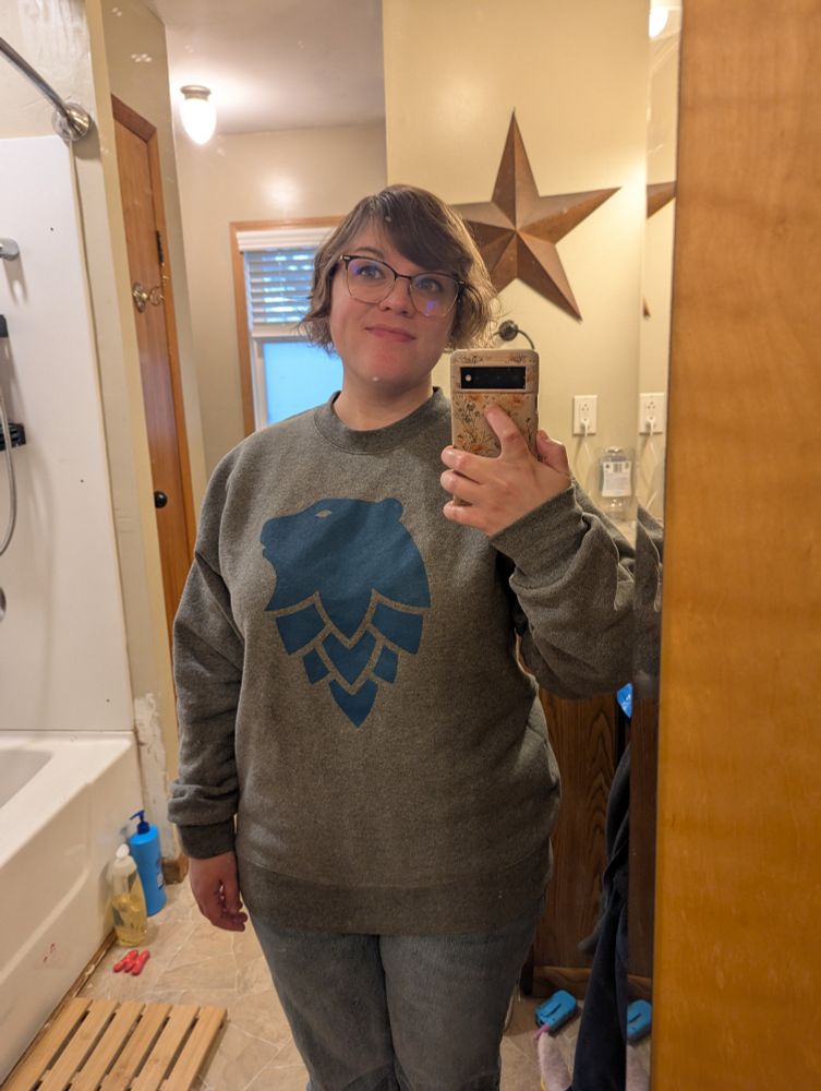 Picture of me taking a selfie in the bathroom mirror. I'm wearing jeans and a grey sweatshirt with a bear head that transitions into some hops. I have short brown hair and black and gold rimmed glasses.